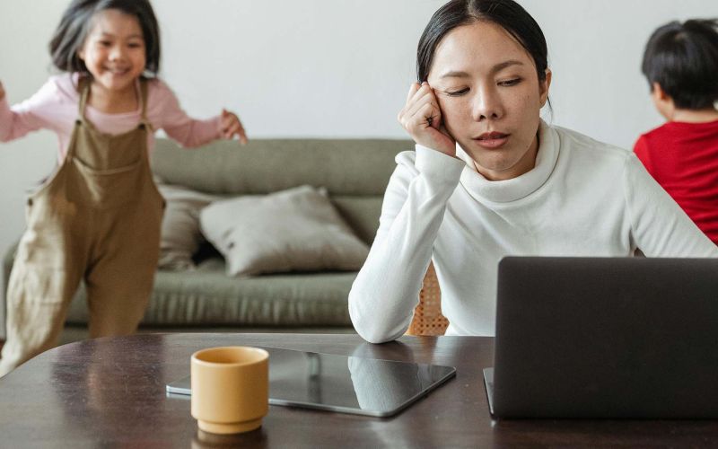 en mamma sitter trött med sin laptop och barnen leker i bakgrunden
